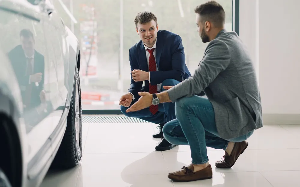 A salesman crouching to talk to a customer beside a car